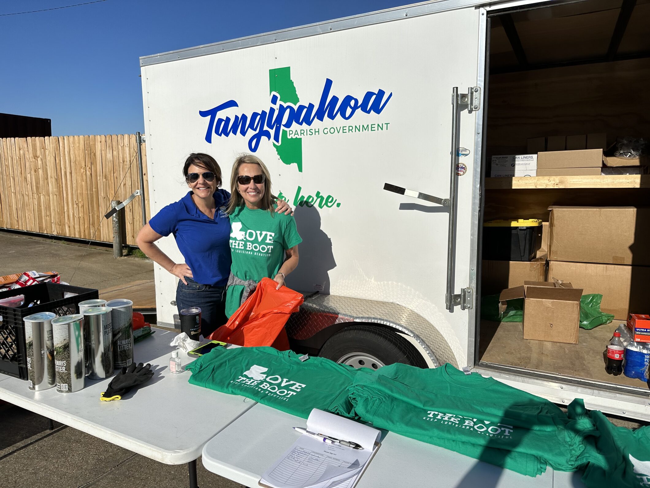 Two women smiling at a volunteer check-in table holding Love the Boot t-shirts and cleanup supplies, standing in front of an open Tangipahoa Parish Government supply trailer.
