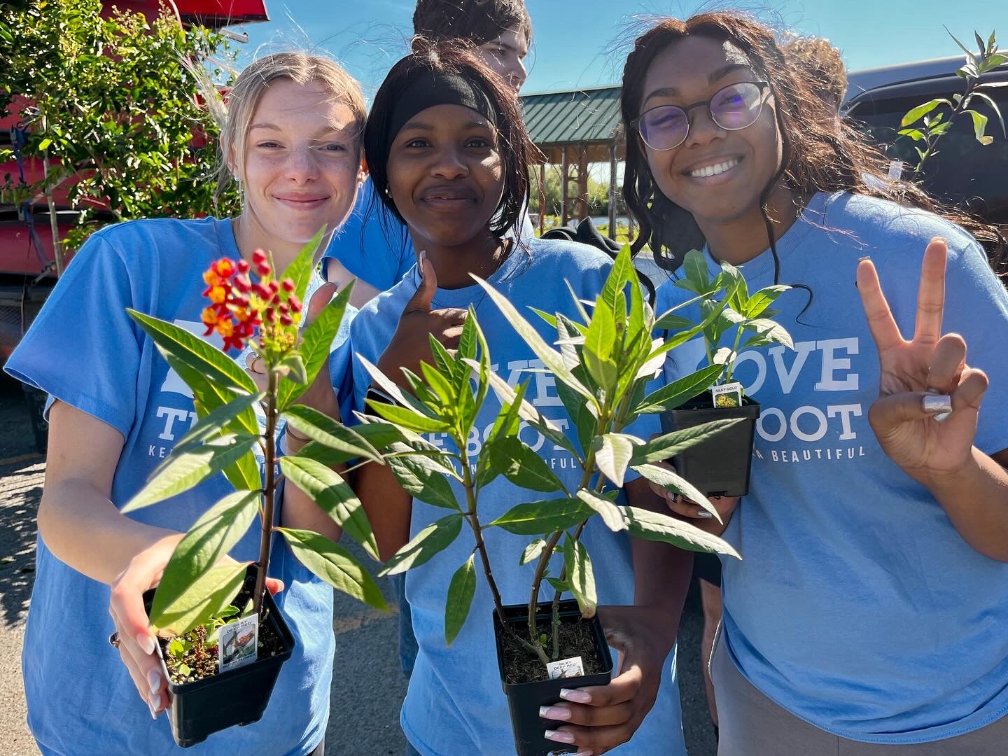 Three young volunteers smiling and holding potted plants while wearing light blue Love the Boot campaign t-shirts.