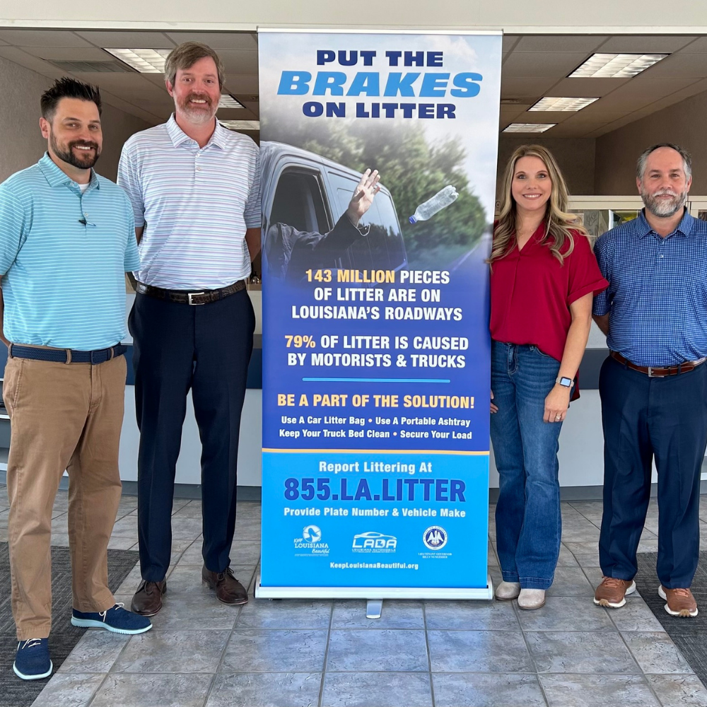 Four people standing in a car dealership next to a sign that says "Put the Brakes on Litter"