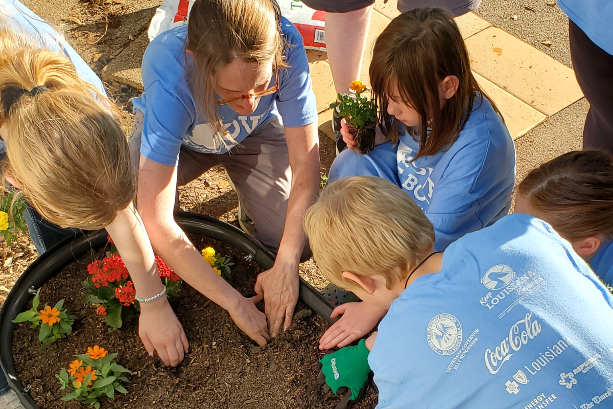 An adult and several children wearing light blue Love the Boot volunteer shirts planting flowers together in a large planter.