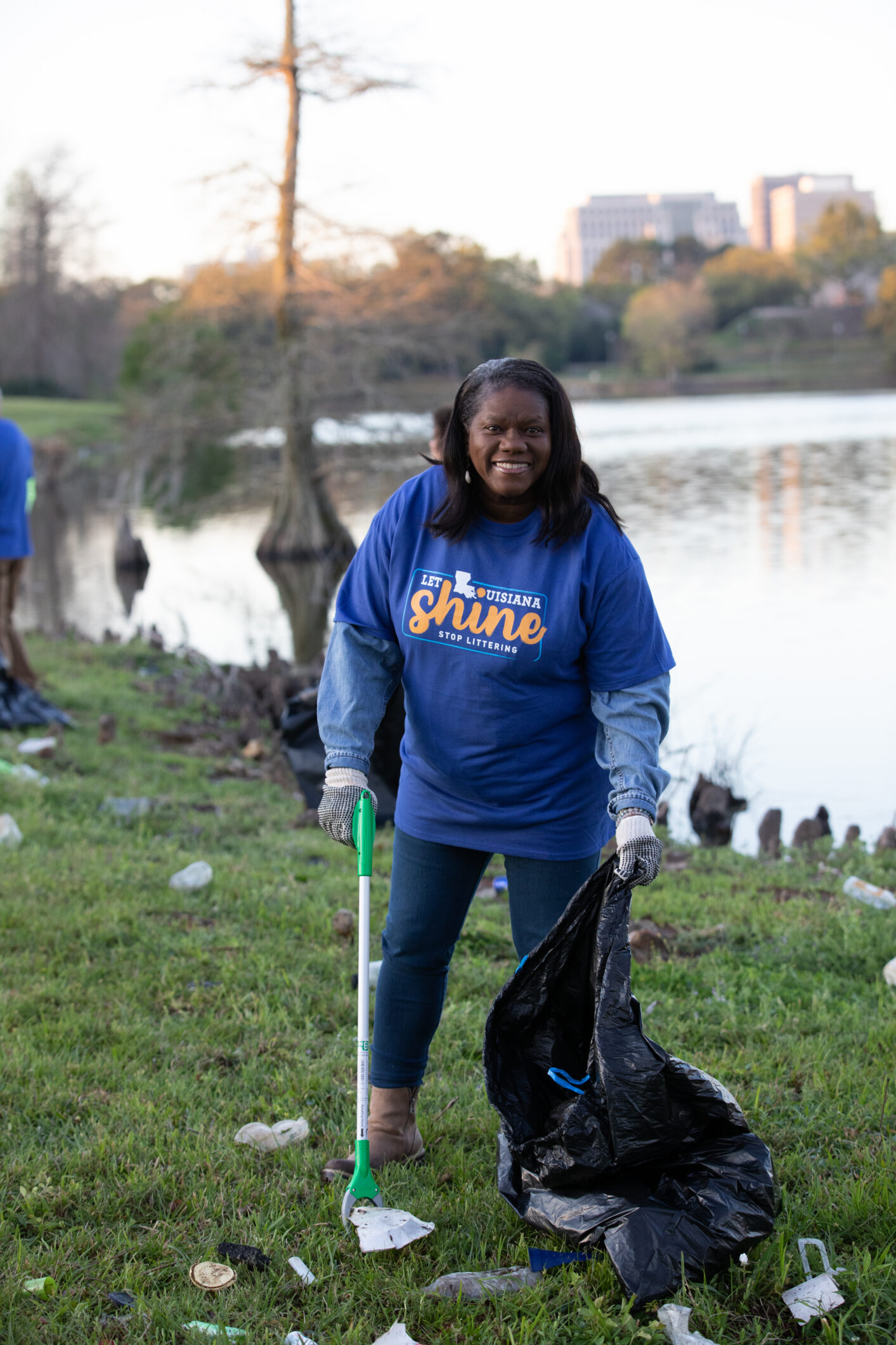 A smiling volunteer in a blue "Let Louisiana Shine, Stop Littering" shirt picks up trash near a lake.