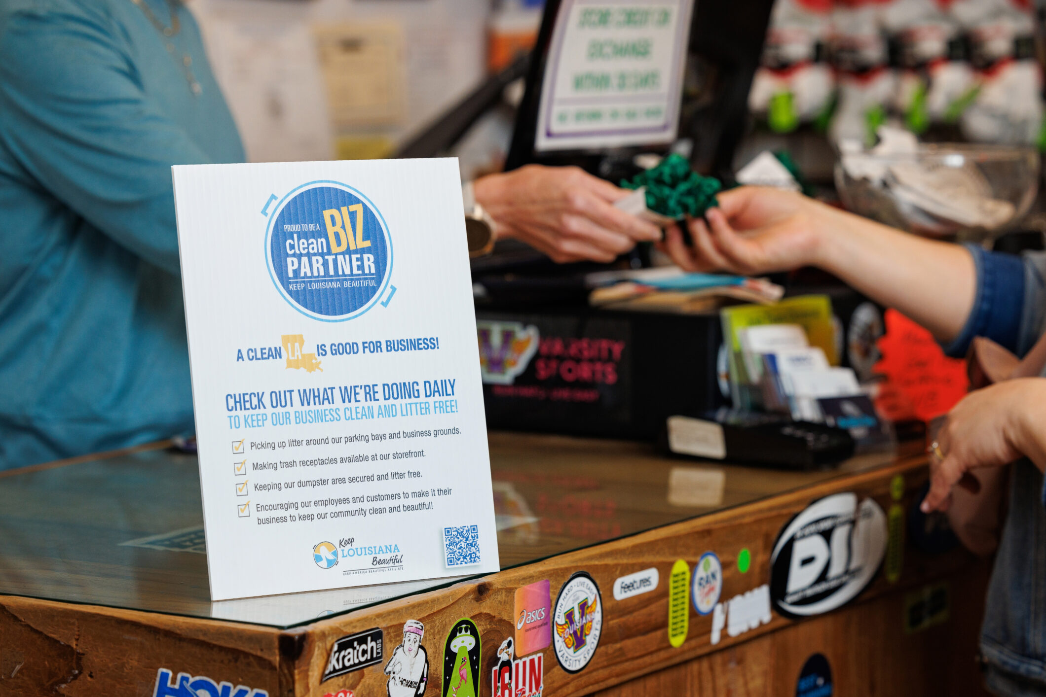 A sign on a checkout counter promoting the Clean Biz Program. Hands exchanging money is in the background behind it.
