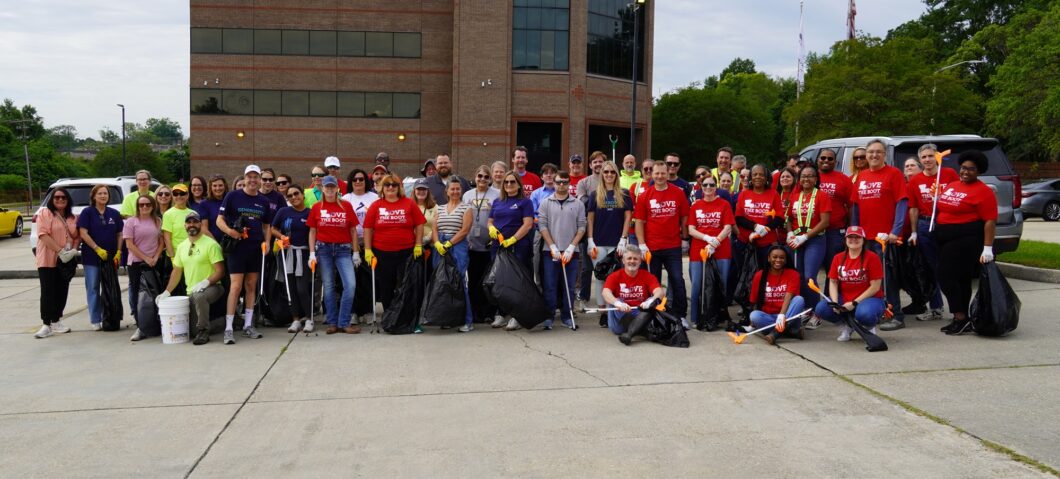 A large group of volunteers posing outdoors during a community cleanup event. Participants are holding trash bags, litter grabbers, buckets, and cleanup supplies in front of an office building parking lot. Many are wearing red Love the Boot Week shirts and smiling for a group photo.