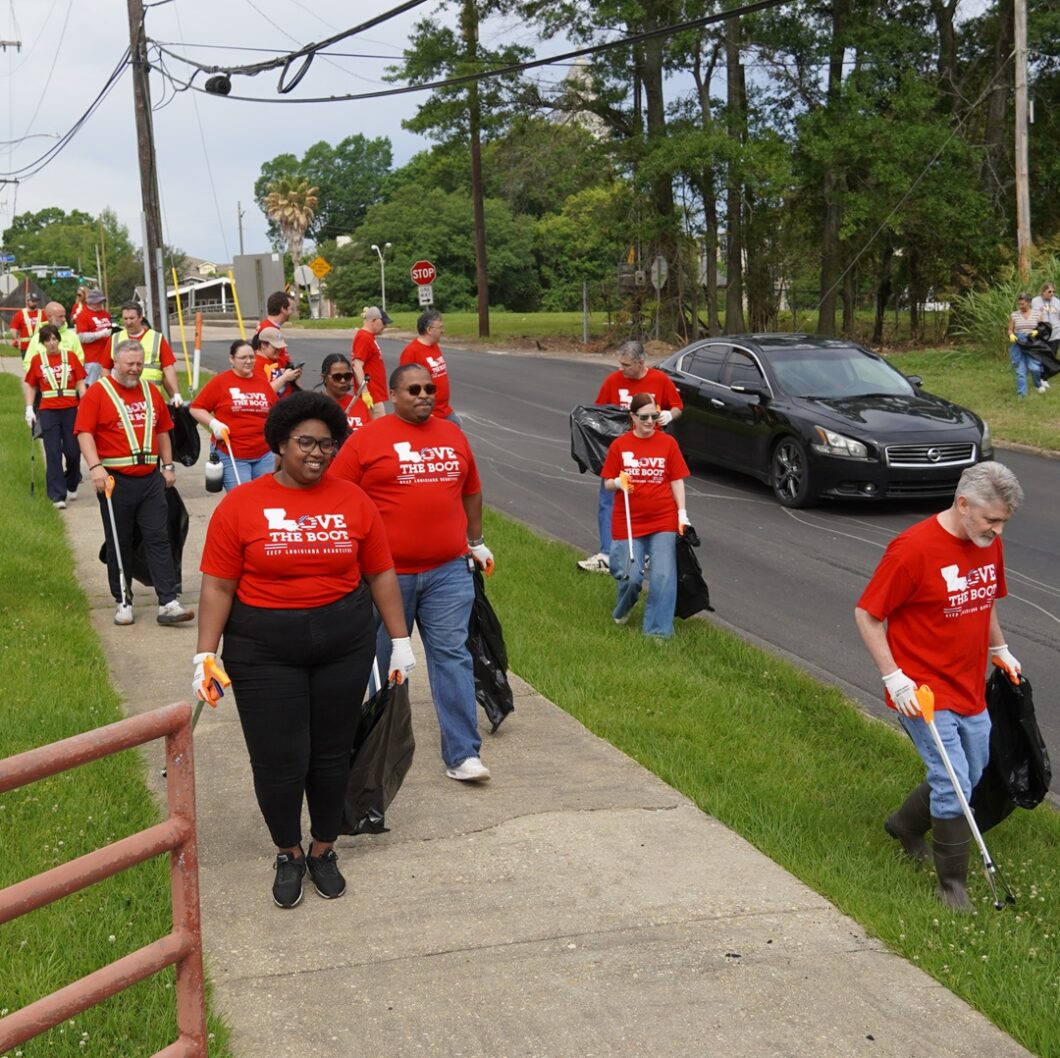 Volunteers wearing red Love the Boot Week shirts walk along a roadside collecting litter with grabbers and trash bags during a community cleanup event.