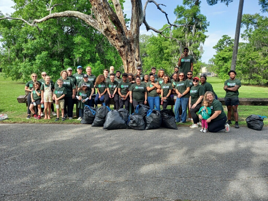 A large group of volunteers in green t-shirts posing together with several full bags of collected litter in a park.