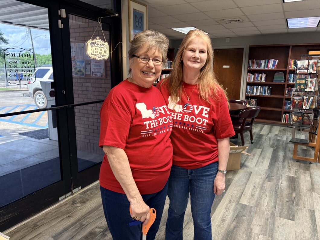 Two volunteers wearing red Love the Boot Week shirts stand inside the Evangeline Parish Library smiling for a photo. One volunteer is holding a litter grabber tool.