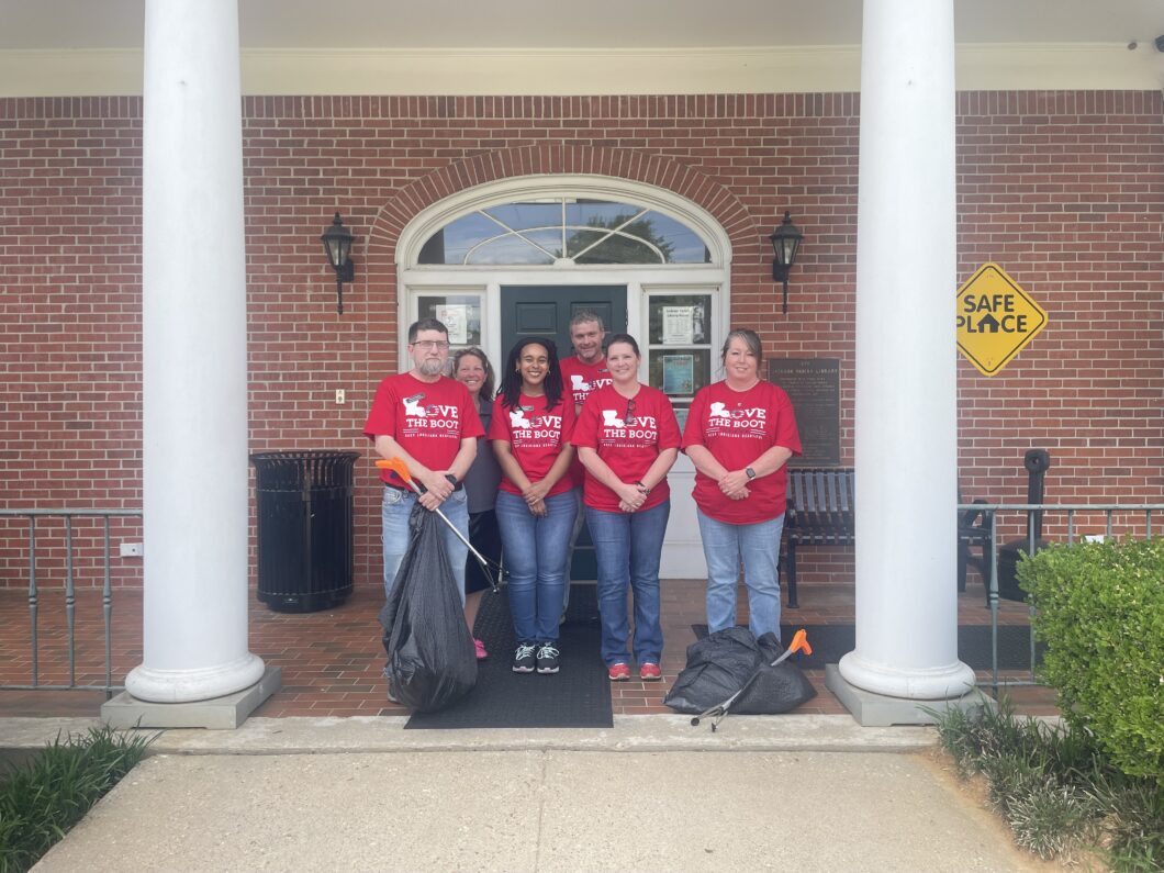 A group of volunteers wearing red Love the Boot Week shirts stand outside Jackson Parish Library holding trash bags and litter grabbers before a cleanup event.
