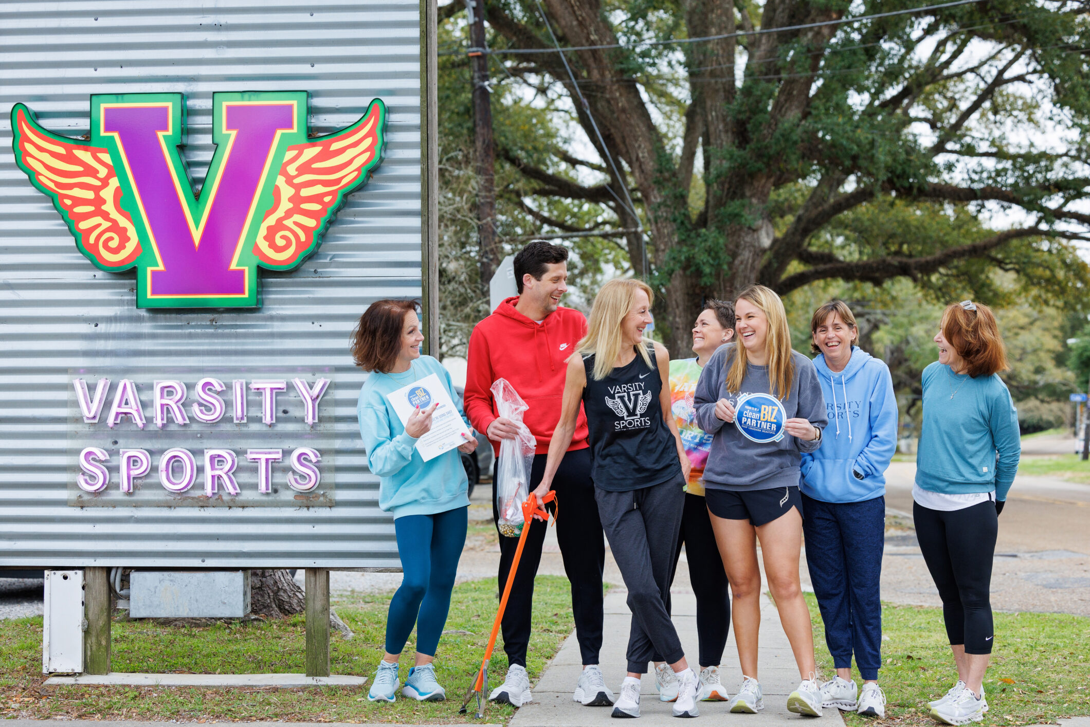 A group of employees standing outside Varsity Sports holding a "Clean Biz Partner" window decal and cleanup tools.