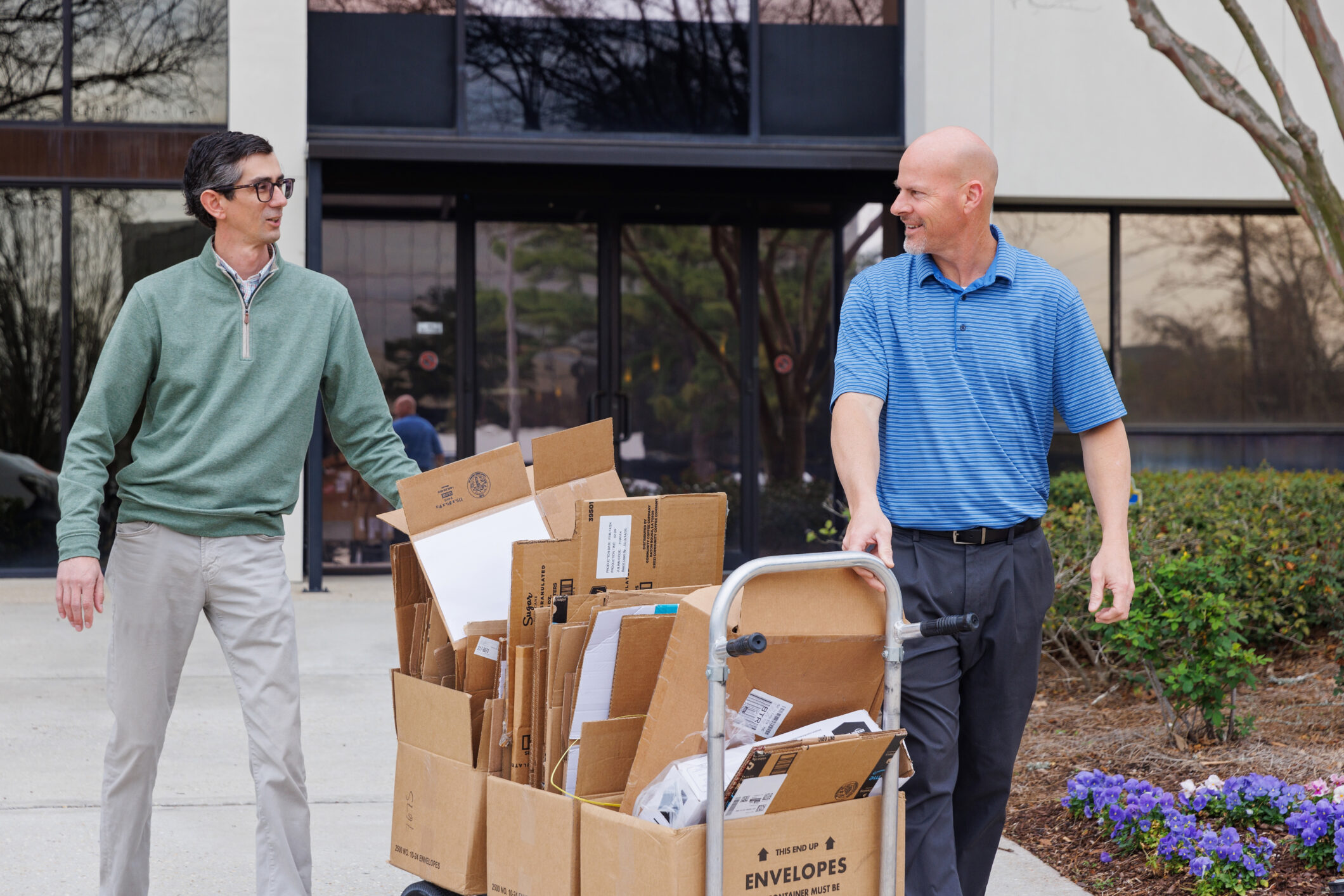 Two men moving a large cart filled with flattened cardboard boxes for recycling outside of an office building.