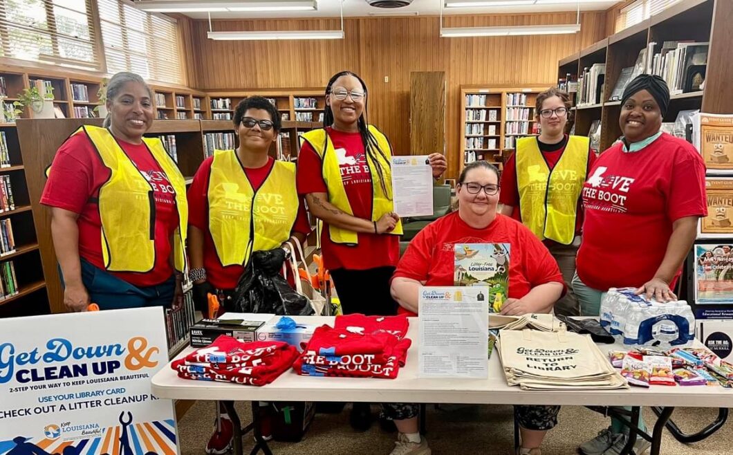 A group of volunteers wearing red Love the Boot Week shirts and yellow safety vests stand behind a table filled with cleanup supplies, tote bags, and informational materials inside Martin Library.