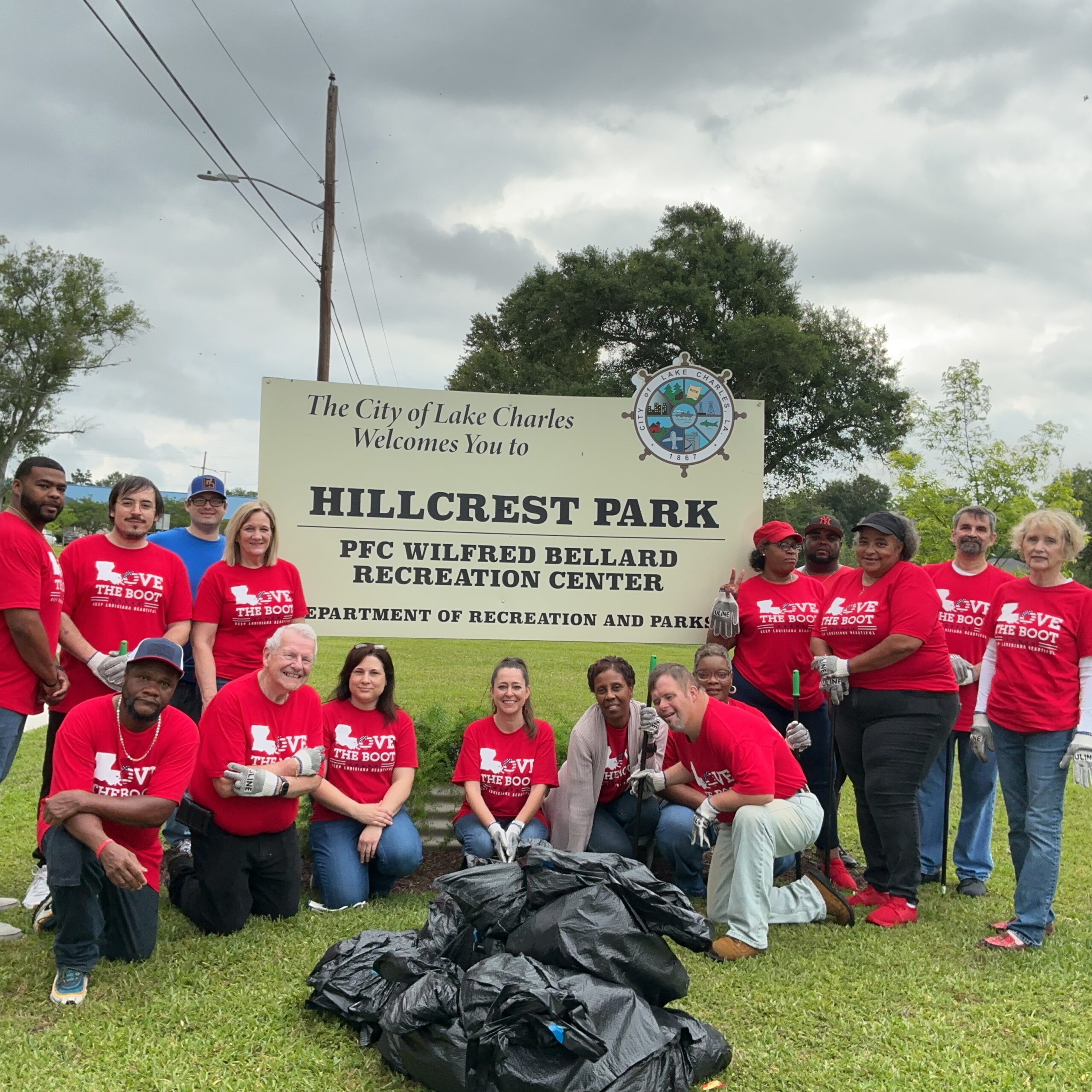 A group of volunteers wearing red Love the Boot Week shirts pose in front of the Hillcrest Park sign in Lake Charles. Several bags of collected litter are displayed in front of the group following a community cleanup event.