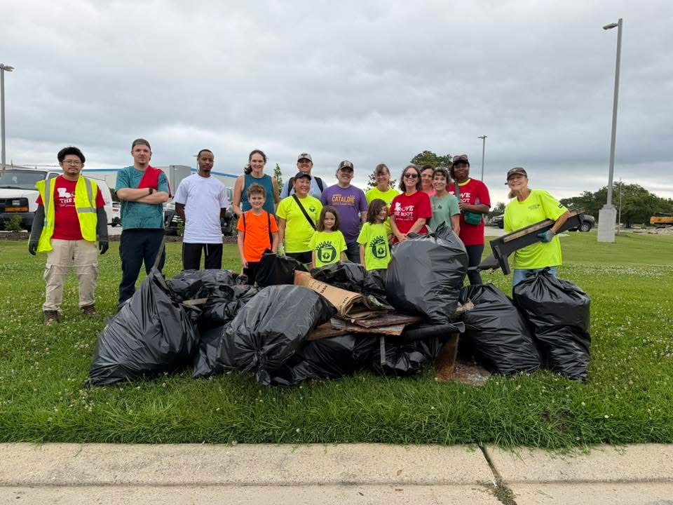 A group of Keep Tiger Town Beautiful volunteers, including adults and children, stand behind a large pile of filled trash bags collected during a Love the Boot Week cleanup event. Volunteers pose on a grassy area with the collected litter in front of them.