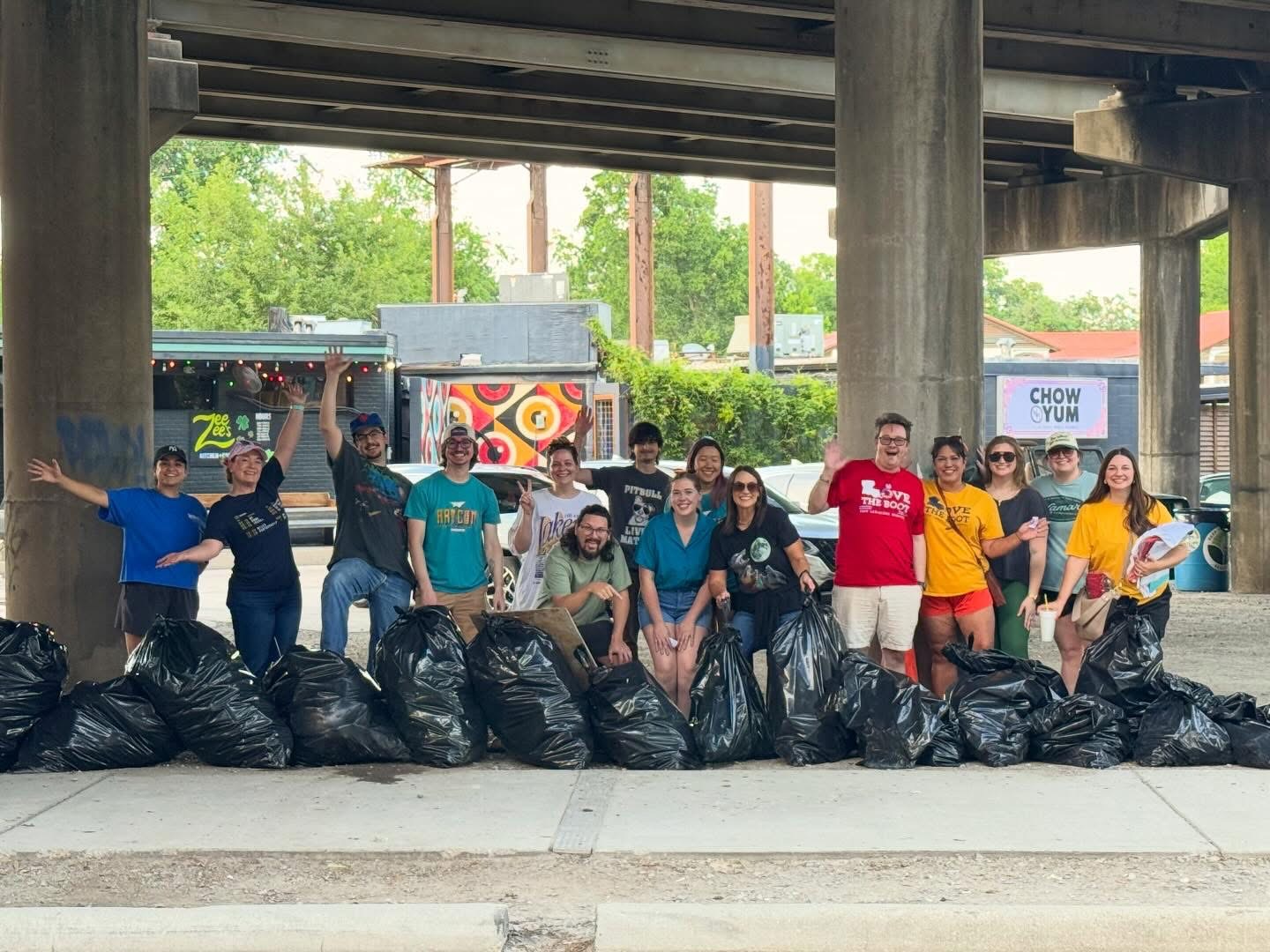 A group of Keep Tiger Town Beautiful volunteers pose under an overpass beside numerous bags of collected litter during a Love the Boot Week cleanup event. Participants smile and wave while standing behind the filled trash bags.