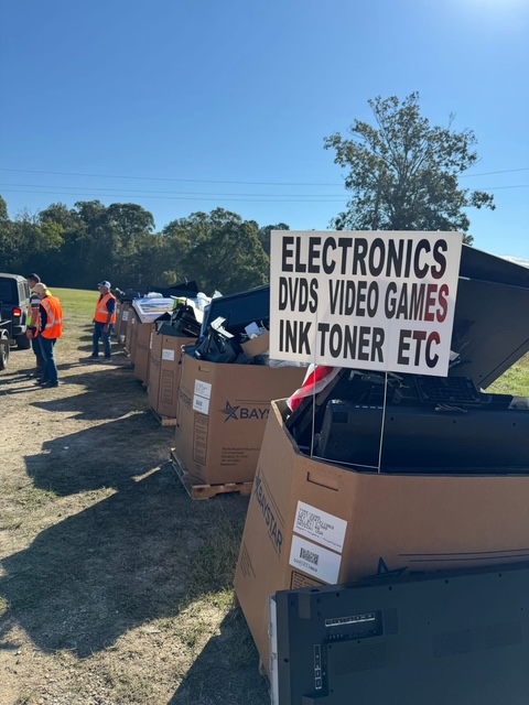 Large collection bins filled with electronics and other recyclable materials are lined up at a drive-through disposal event hosted by Livingston Parish and the Livingston Parish Sheriff’s Office. A sign indicates accepted items, including electronics, DVDs, video games, and ink toner.