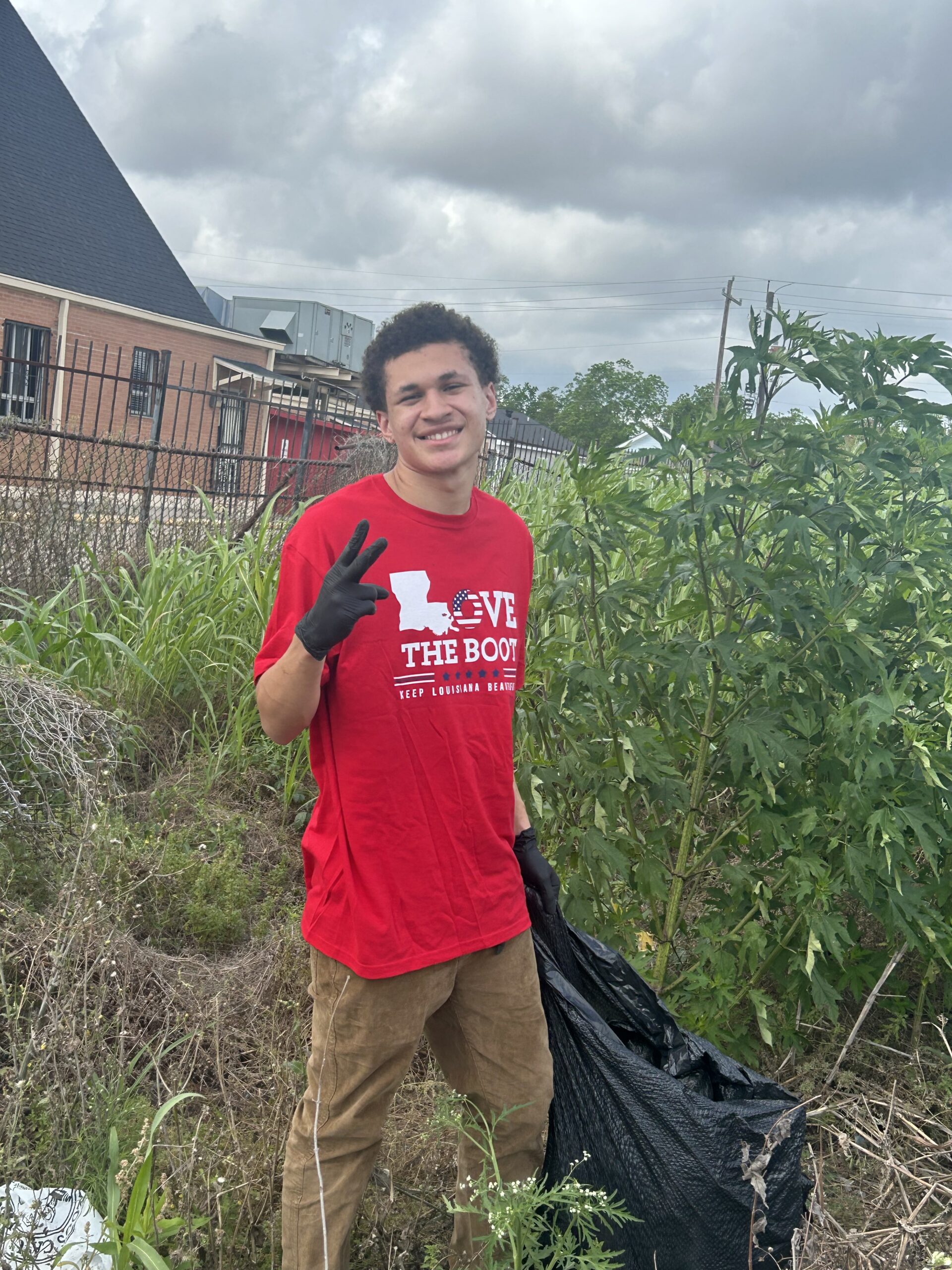 A student wearing a red Love the Boot Week shirt and black gloves stands outdoors holding a trash bag during a community cleanup event. The student smiles and makes a peace sign while volunteering in a grassy area near a neighborhood.