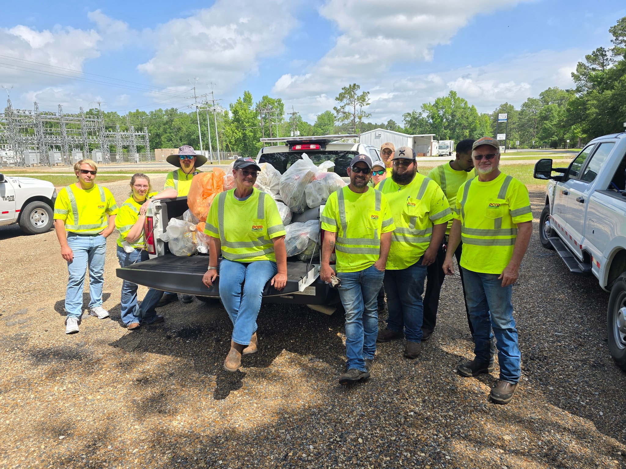 A group of Louisiana Department of Transportation and Development (DOTD) employees wearing high-visibility safety shirts stand around a pickup truck filled with bags of collected litter during a Love the Boot Week cleanup event.