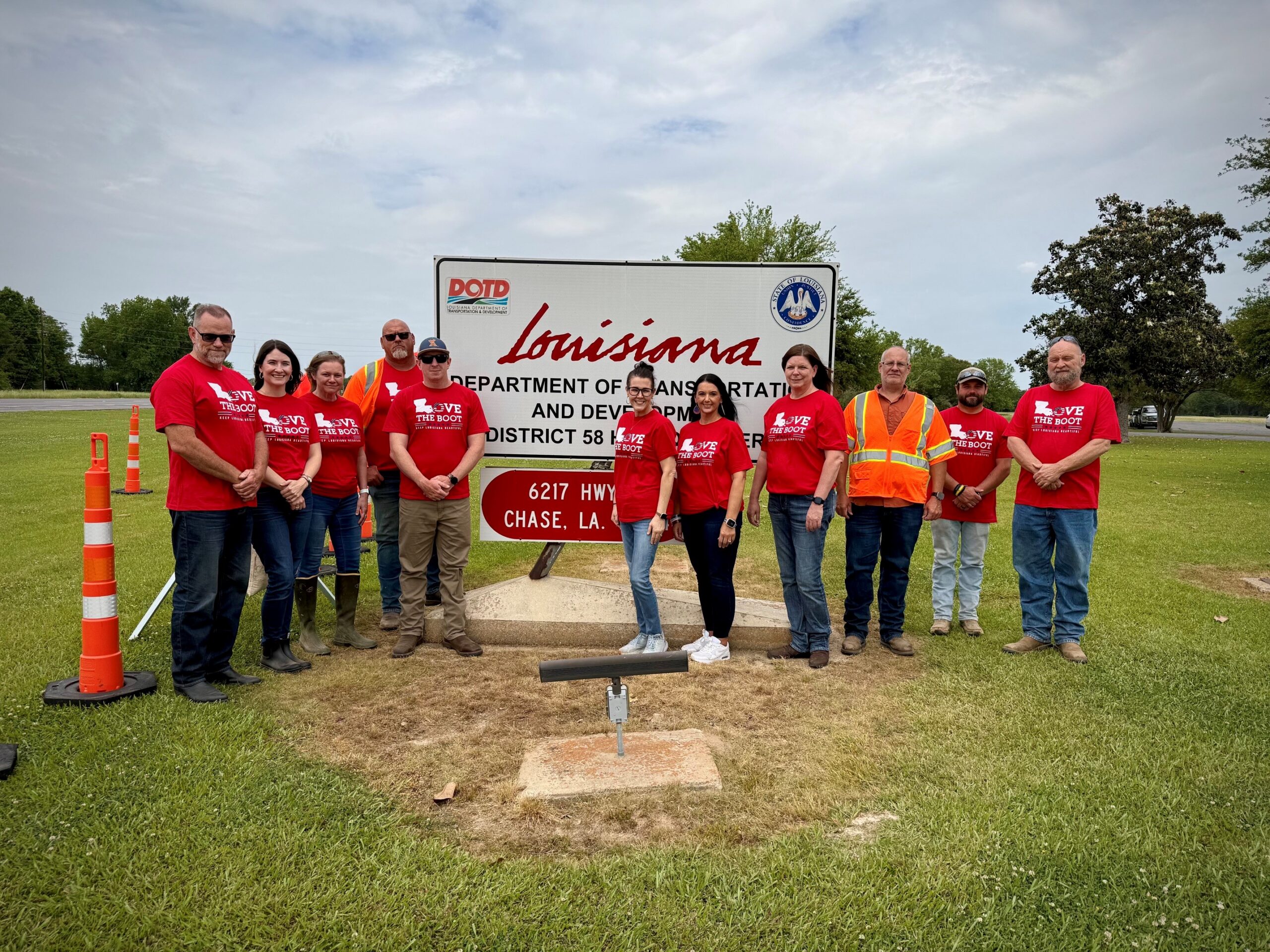 A group of Louisiana Department of Transportation and Development (DOTD) employees and volunteers wearing red Love the Boot Week shirts and safety vests stand in front of a DOTD district office sign during a community cleanup event.