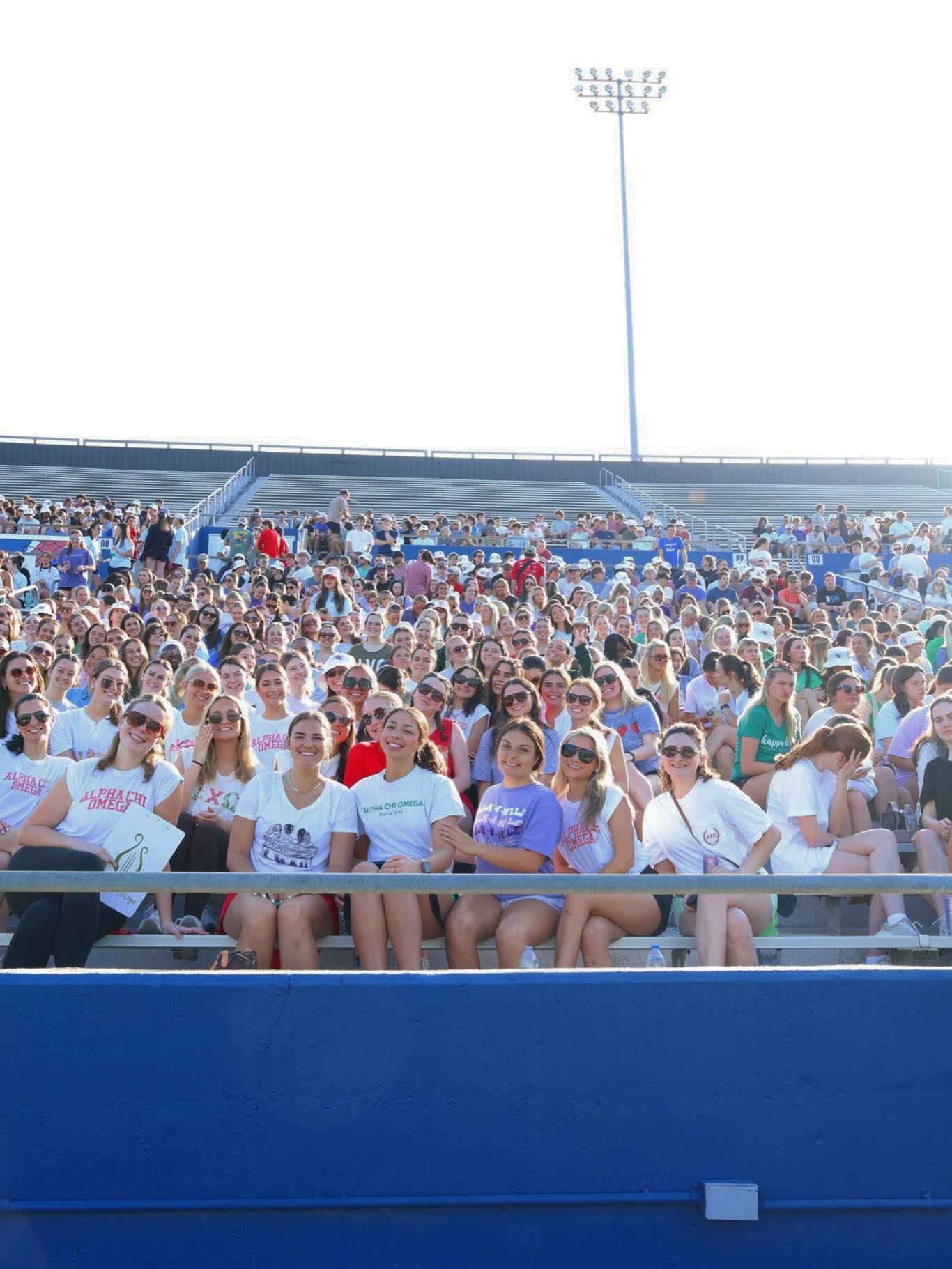 A large group of college students sit together in stadium bleachers during a campus event. Many participants are wearing matching student organization shirts and smiling for a group photo in the stands.