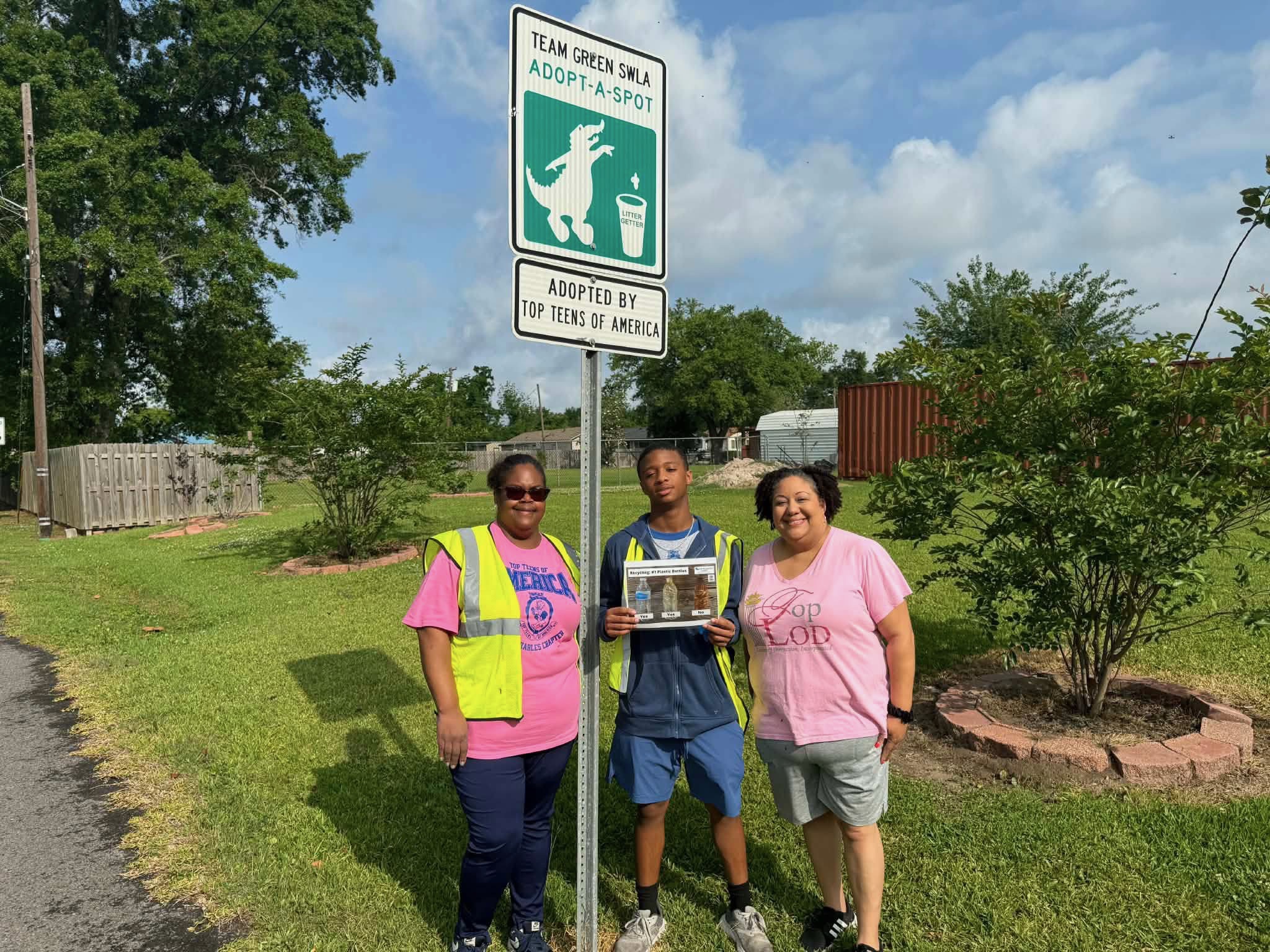 Three volunteers stand beside a Team Green SWLA Adopt-A-Spot sign in Lake Charles. One participant holds an educational display while the group poses near a maintained roadside area during a Love the Boot Week community improvement project.