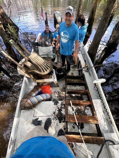 Volunteers in boats remove large debris, including a chair and ladder, from a wooded waterway during a Love Our Waterways cleanup.