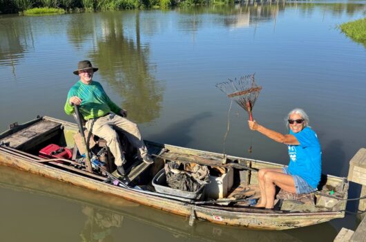Two volunteers in a small boat collect litter and debris from a waterway using cleanup tools.