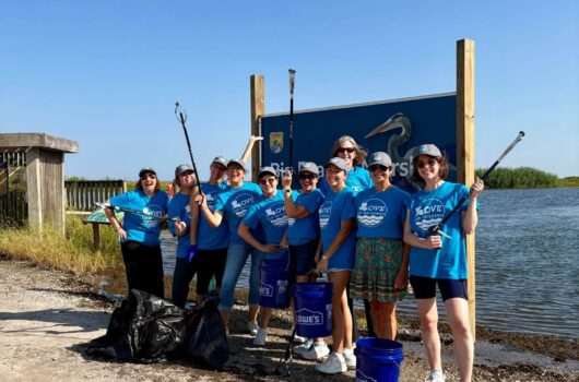 Group of Love Our Waterways volunteers pose with buckets, litter grabbers and collected trash beside a coastal waterway.