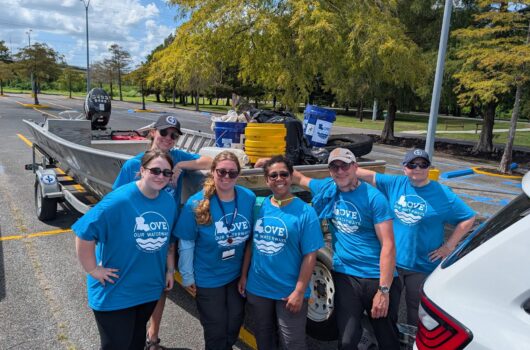Five volunteers wearing blue Love Our Waterways T-shirts pose for a group photo in a parking lot beside a boat and trailer filled with cleanup supplies.